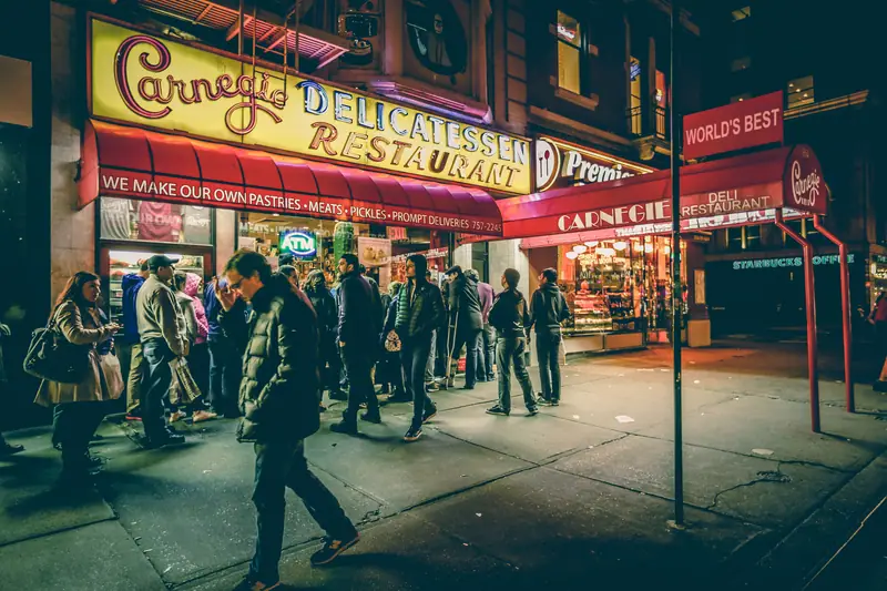 A busy New York street corner with a lit-up deli at night